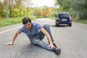 Man sitting in road with injured ankle after struck by car, with car driving away in the background in hit-and-run.