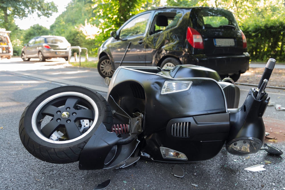 Damaged scooter lying on the road after a collision with a car, with visible vehicle damage in the background.